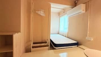 A compact room with a bed, a small table, a chair, a wall-mounted air conditioner, and a window with curtains.