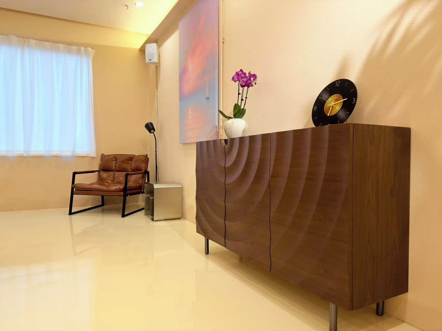 A modern room with a wooden cabinet, a leather chair, a vase with flowers, and a wall-mounted clock.