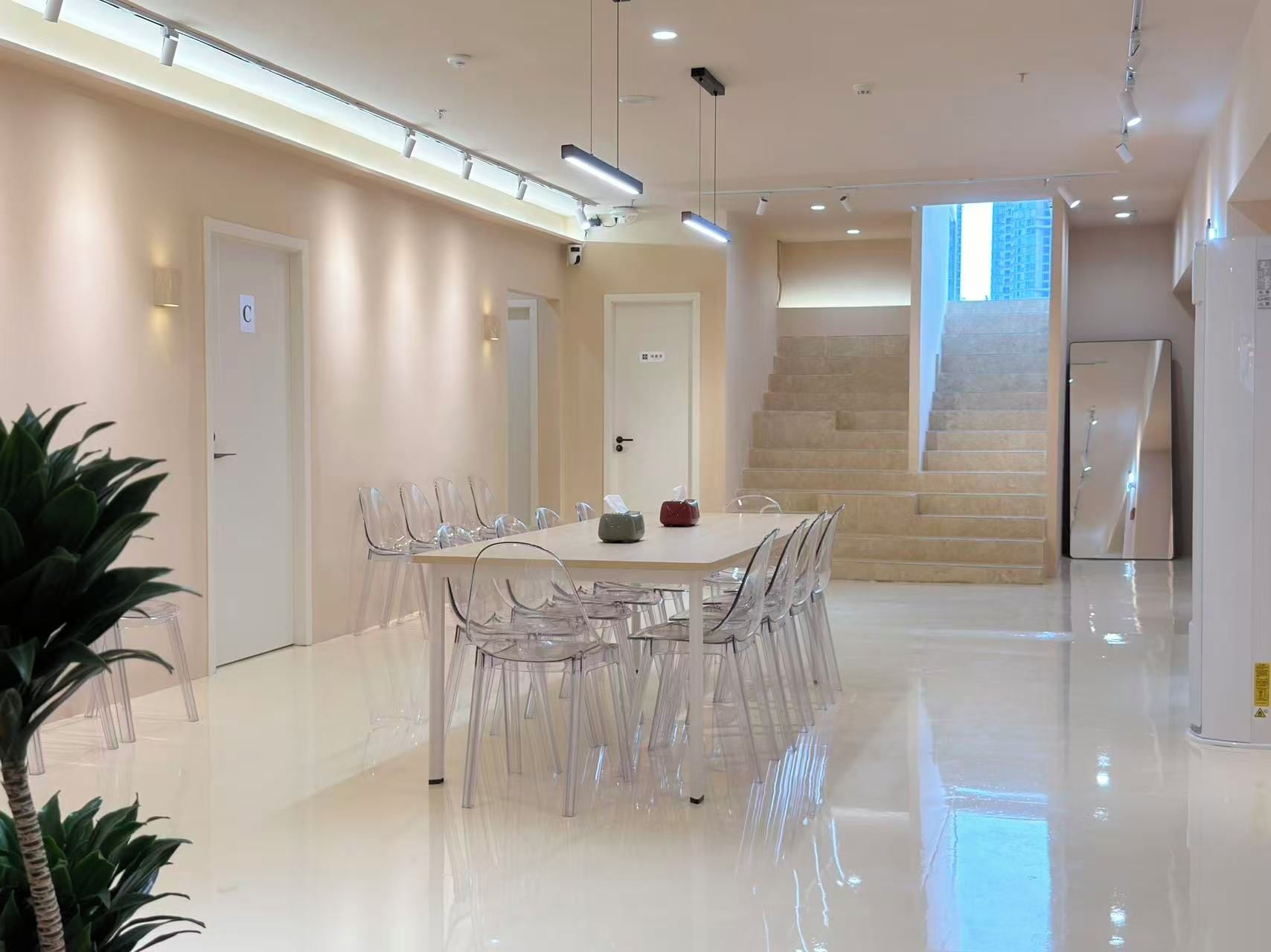 A modern hotel room with a large dining table, clear chairs, a staircase, and a large window.