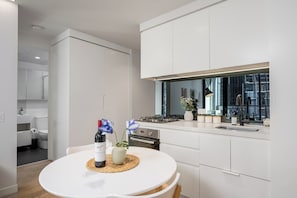 A modern kitchen with white cabinetry, a round dining table with a placemat, and a bottle of wine. - Central Melbourne 2BR - Pool, Gym & Free Trams (Melbourne)