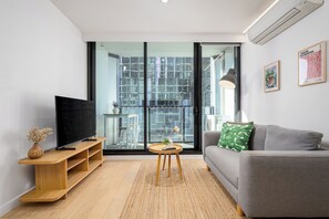 A modern living room with a flat-screen TV, a grey sofa, a small round table, and a large glass door leading to an outdoor balcony. - Central Melbourne 2BR - Pool, Gym & Free Trams (Melbourne)
