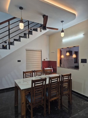 A dining area with a marble table and wooden chairs, a staircase with a black railing, and pendant lighting. - Serene Green Retreat Pothencode (Thiruvananthapuram)