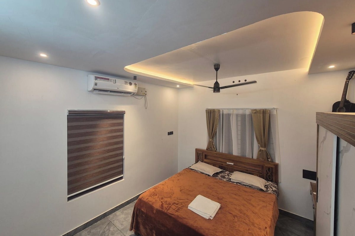 A bedroom with a bed, a ceiling fan, an air conditioning unit, a window with blinds, and a guitar on a stand.