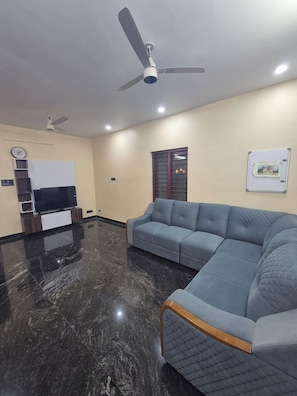 A modern living room with a sectional sofa, a flat-screen TV, ceiling fans, and a marble floor. - Serene Green Retreat Pothencode (Thiruvananthapuram)