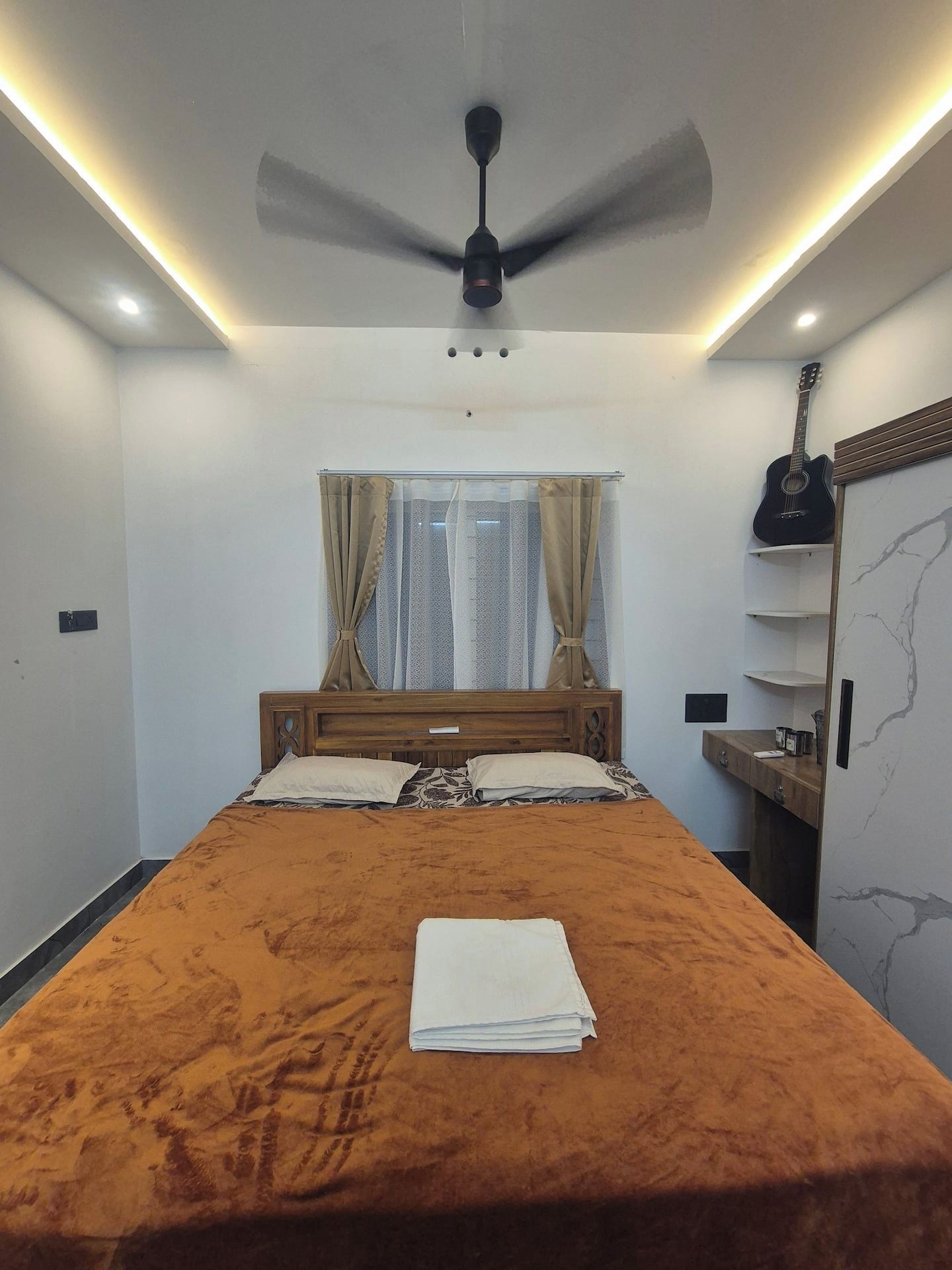 A bedroom with a bed, a ceiling fan, a window with curtains, a guitar, and a marble wall.