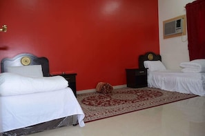 Ein Zimmer mit zwei Betten, einer roten Wand, einem gemusterten Teppich und einem kleinen Fenster mit Klimaanlage.