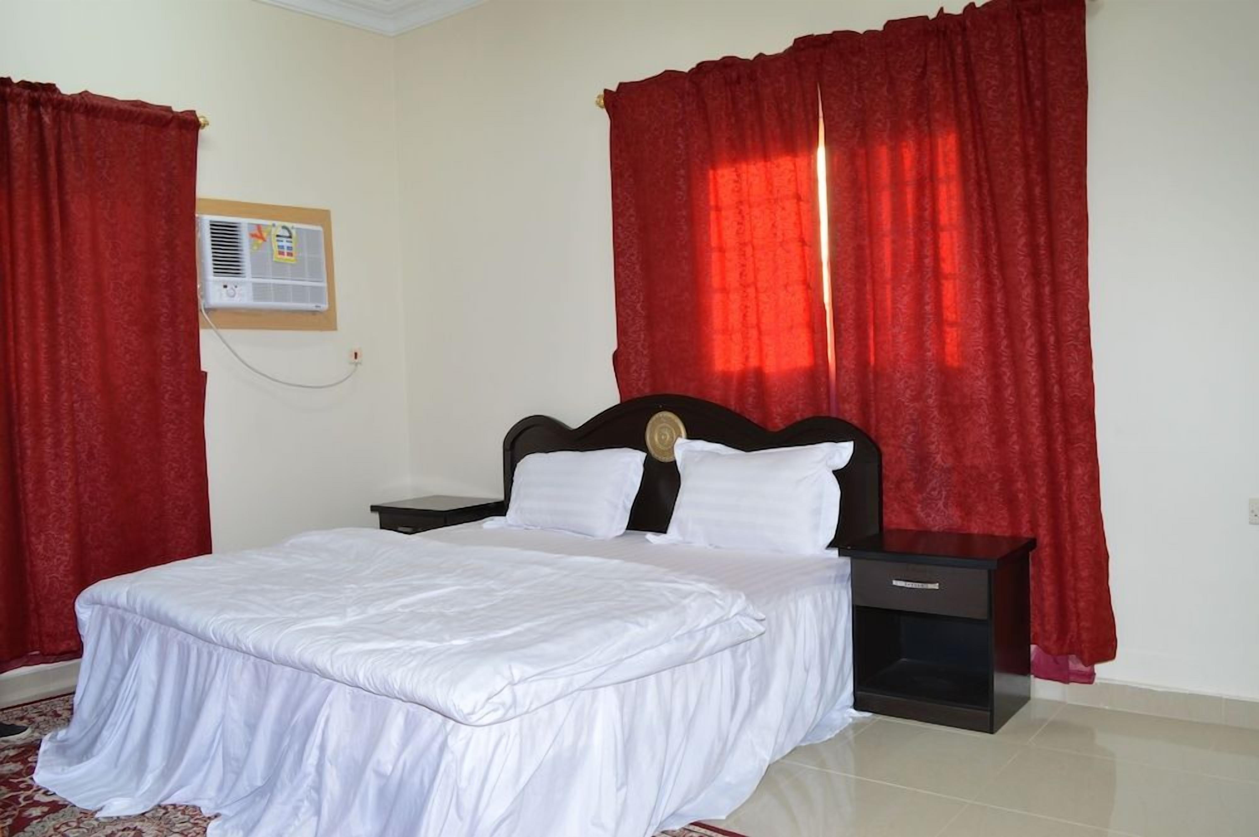 A bedroom with a bed, nightstands, a red curtain, a wall-mounted air conditioner, and a red carpet.