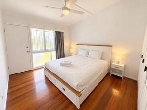 Un dormitorio con cama, ventilador de techo, mesita de noche con lámpara, ventana con cortinas y puerta.