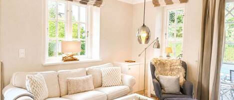 Una sala de estar con sofá, sillón y mesa de centro, ventanas amplias con cortinas y una lámpara.