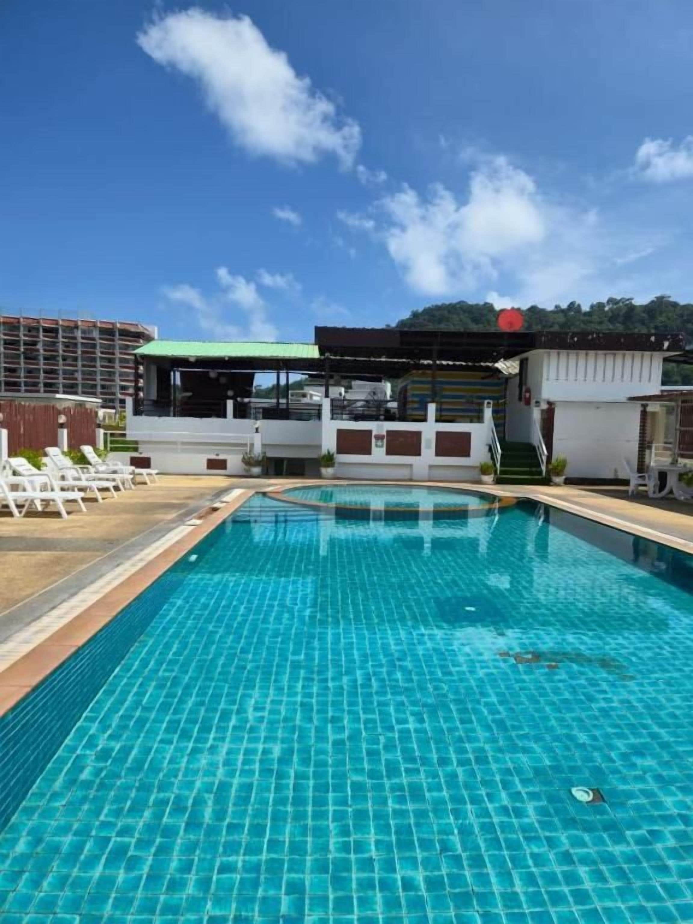 A swimming pool with a tiled surface, surrounded by lounge chairs and a covered area with a green roof.