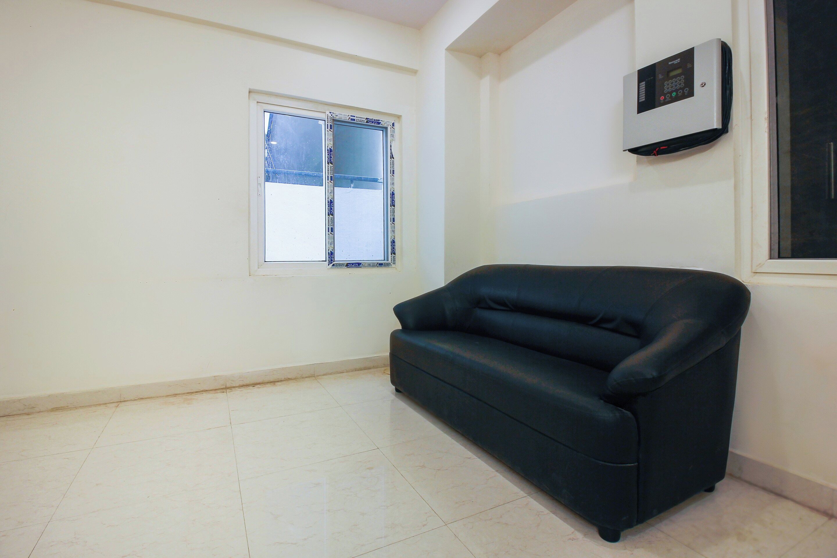A room with a white wall, a window with blinds, a black sofa, and a thermostat on the wall.