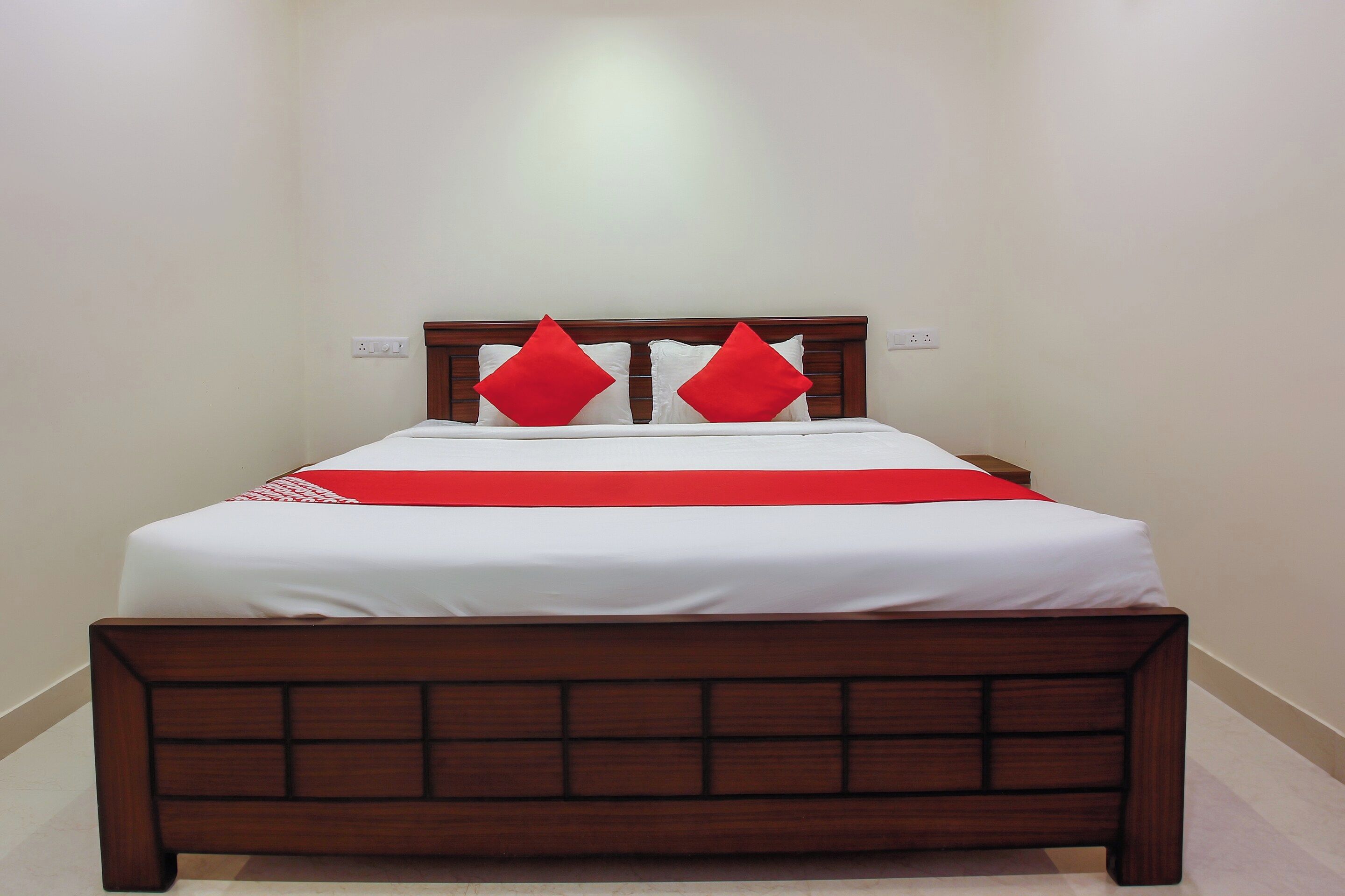 Cama de solteiro com cabeceira de madeira e duas almofadas vermelhas, diante de uma parede lisa com um interruptor de luz.