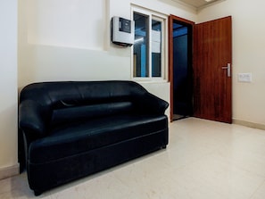 Um quarto com sofá preto, janela, porta de madeira e ar-condicionado fixado na parede.