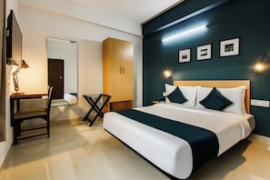 A hotel room with a bed, a desk with a lamp, a wardrobe, and a wall with framed pictures.