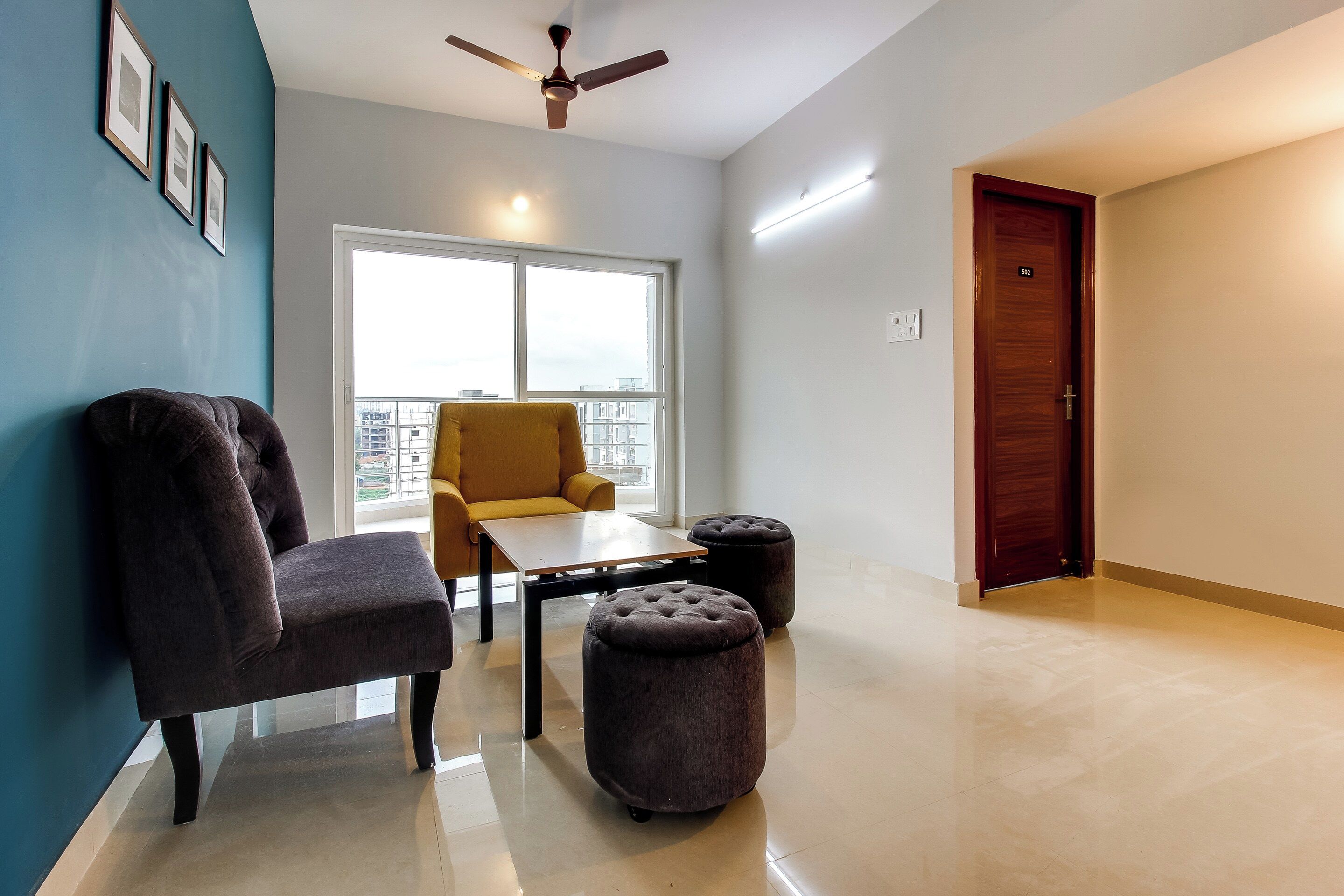 A hotel room with a ceiling fan, a window with a city view, a sofa, a small table, and a door.