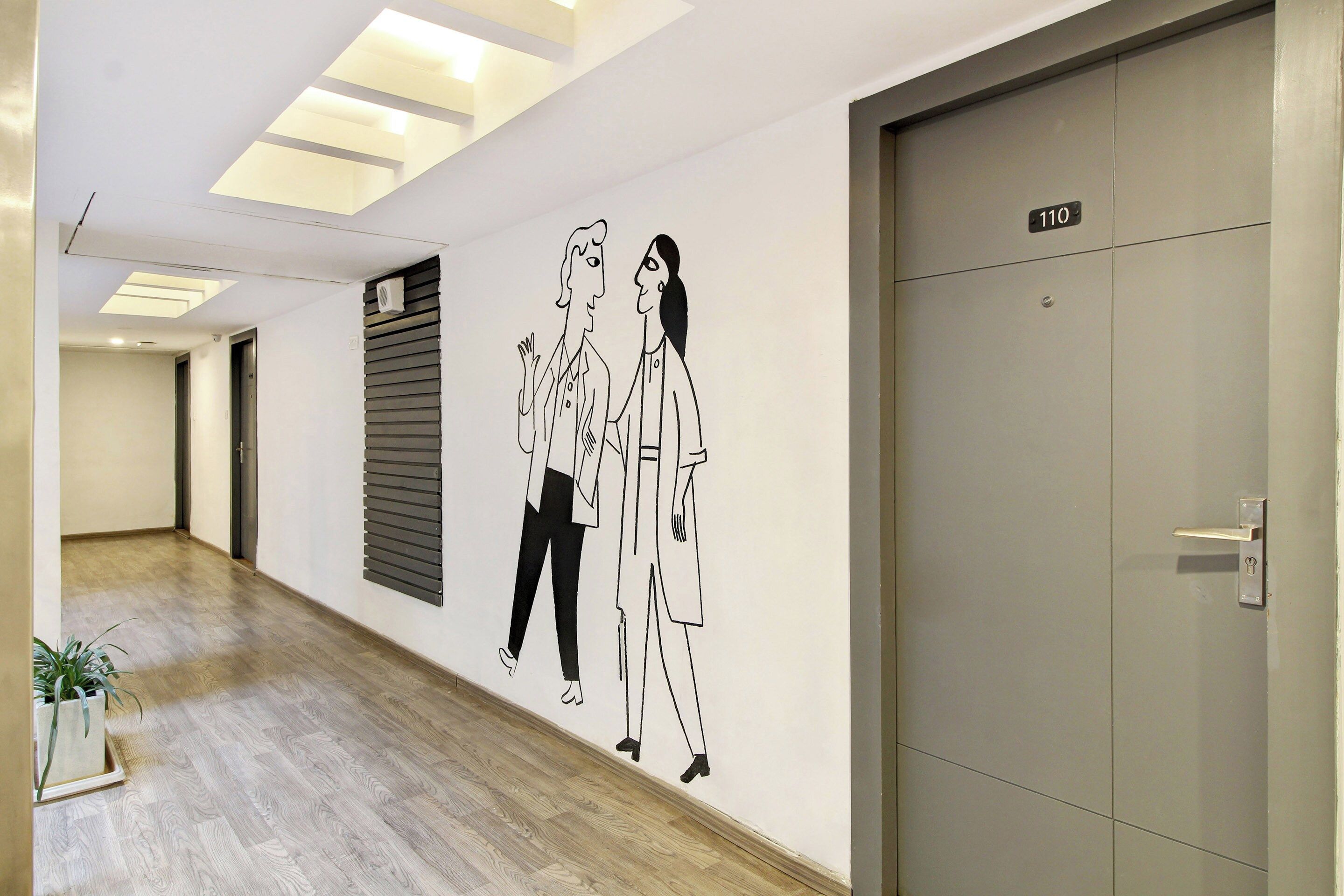 Un pasillo con piso de madera, un equipo de aire acondicionado montado en la pared, una puerta con placa de número y una planta en maceta.