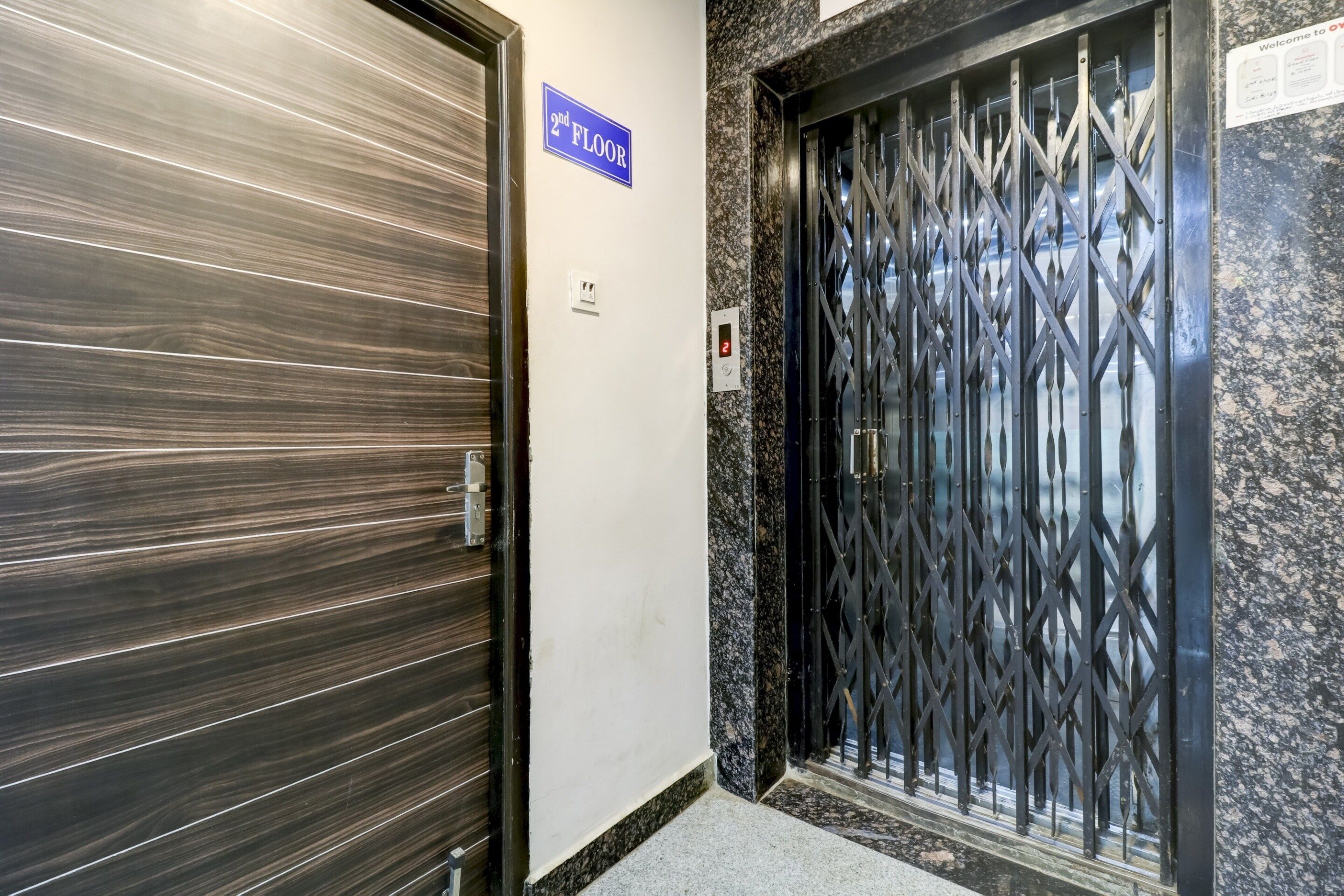 Puerta cerrada de ascensor con un letrero que indica '2do piso', una verja metálica con teclado numérico y una pared con un letrero que dice 'Bienvenidos a nuestro hotel'.