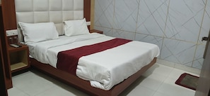 A neatly made bed with white linens and a red accent, a wooden nightstand with a remote control, and a wall with a geometric pattern. - Hotel O R D International (Amritsar)