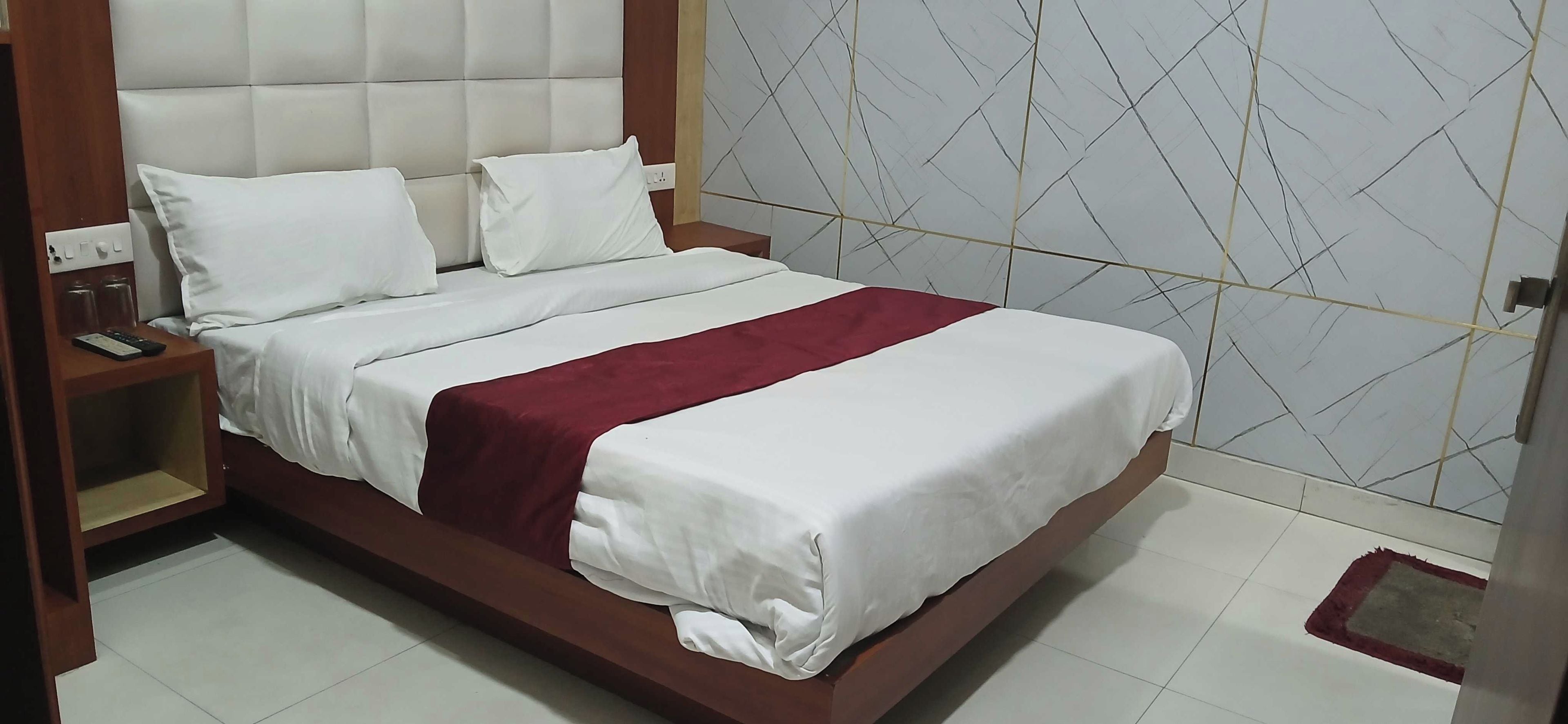 A neatly made bed with white linens and a red accent, a wooden nightstand with a remote control, and a wall with a geometric pattern.