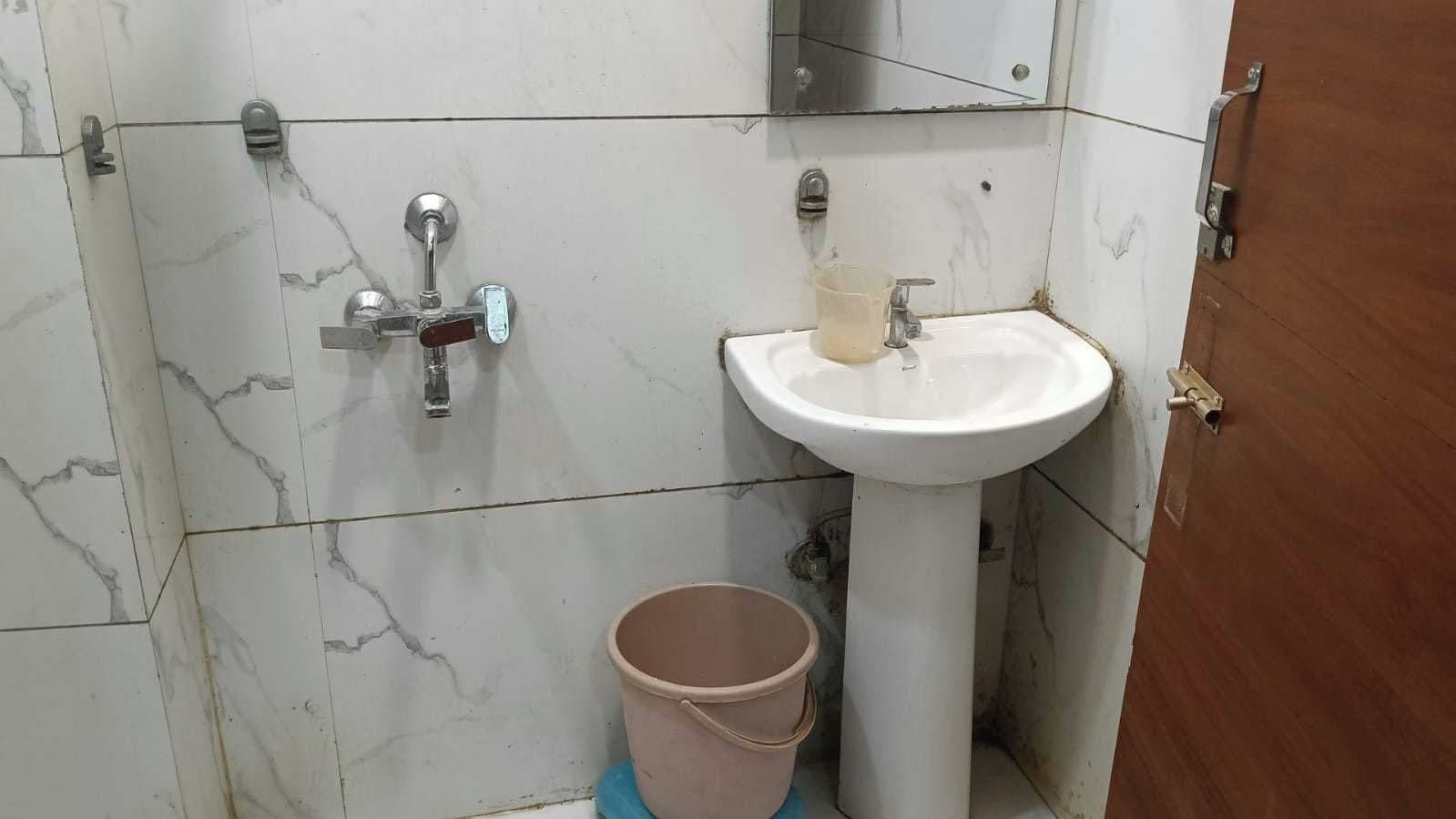 A bathroom with a white sink, a mirror, and a shower with a faucet. There is a bucket on the floor.