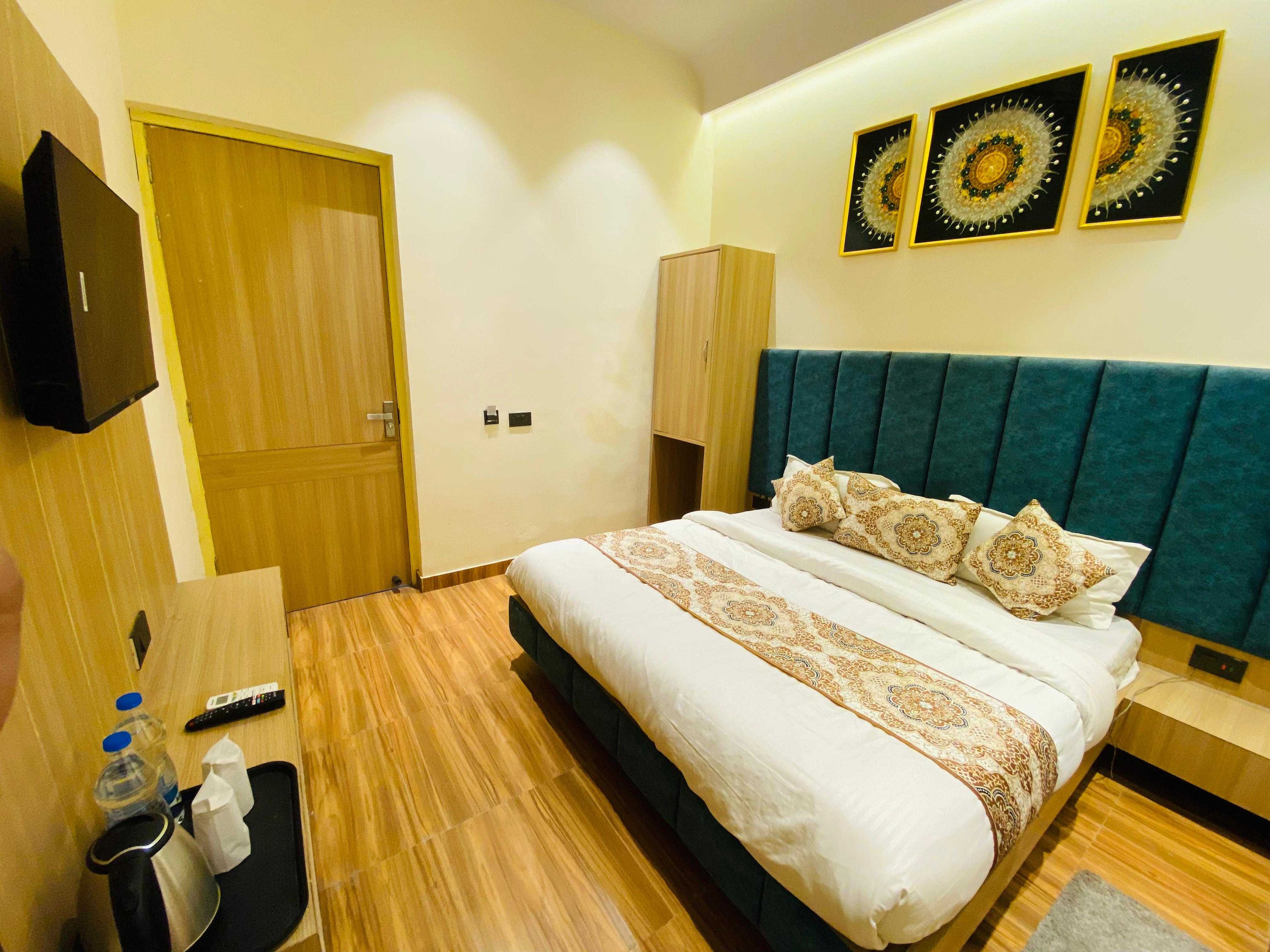 A hotel room with a bed, a TV, a wooden wardrobe, and a small table with a remote control and a bottle.