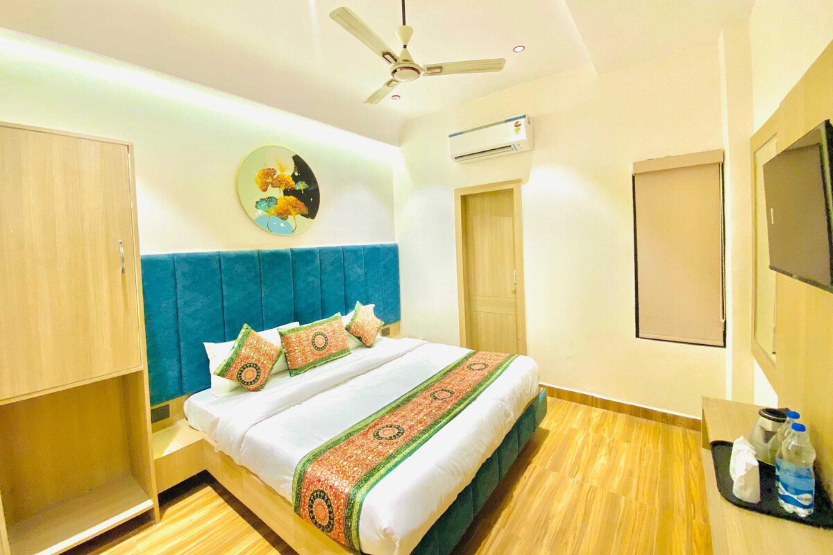 A hotel room with a bed, a ceiling fan, an air conditioning unit, a wall-mounted TV, and a wooden floor.