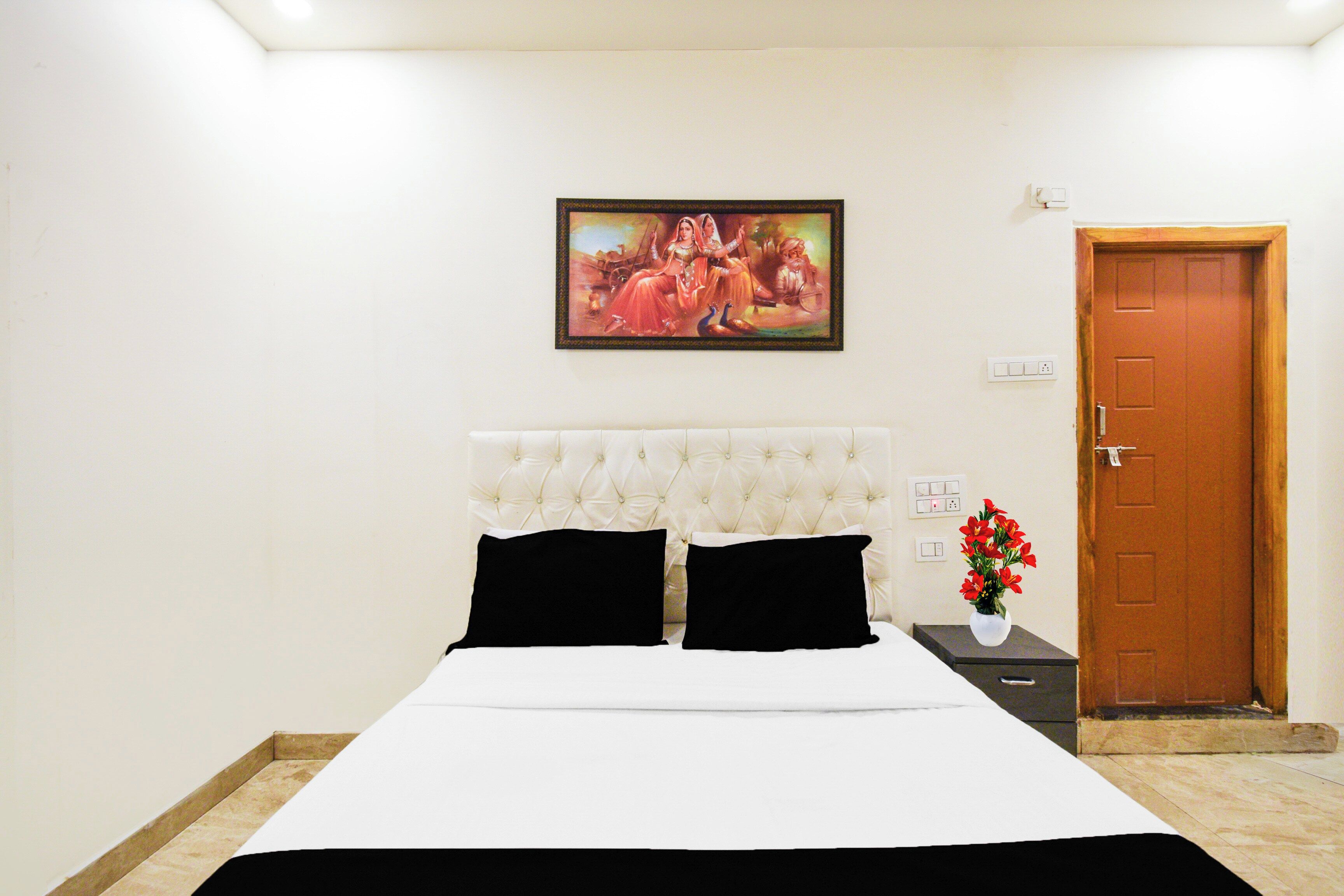 A hotel room with a bed, a painting on the wall, a door, and a nightstand with a vase of flowers.