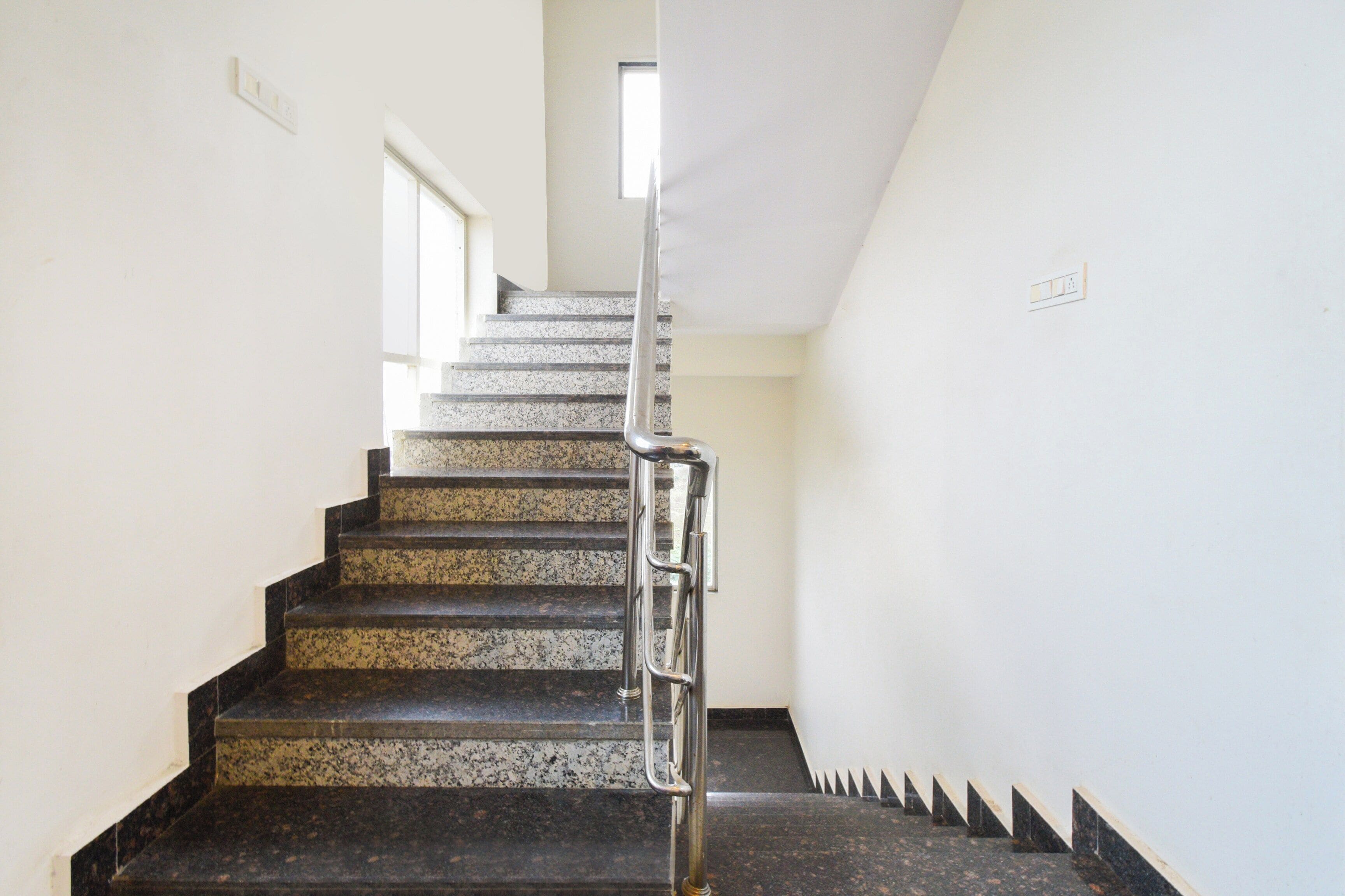 Una escalera con barandilla metálica, peldaños alfombrados y una pared blanca con un ventanal en la parte superior.