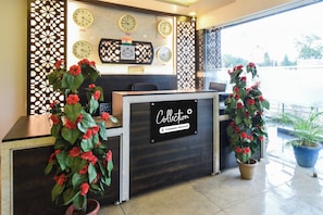 A reception desk with a sign that reads 'Collection Company-Serviced'. There are clocks above the desk displaying different time zones, and two potted plants with red flowers on either side. - Hotel O Charminar international Hotel (Hyderabad)