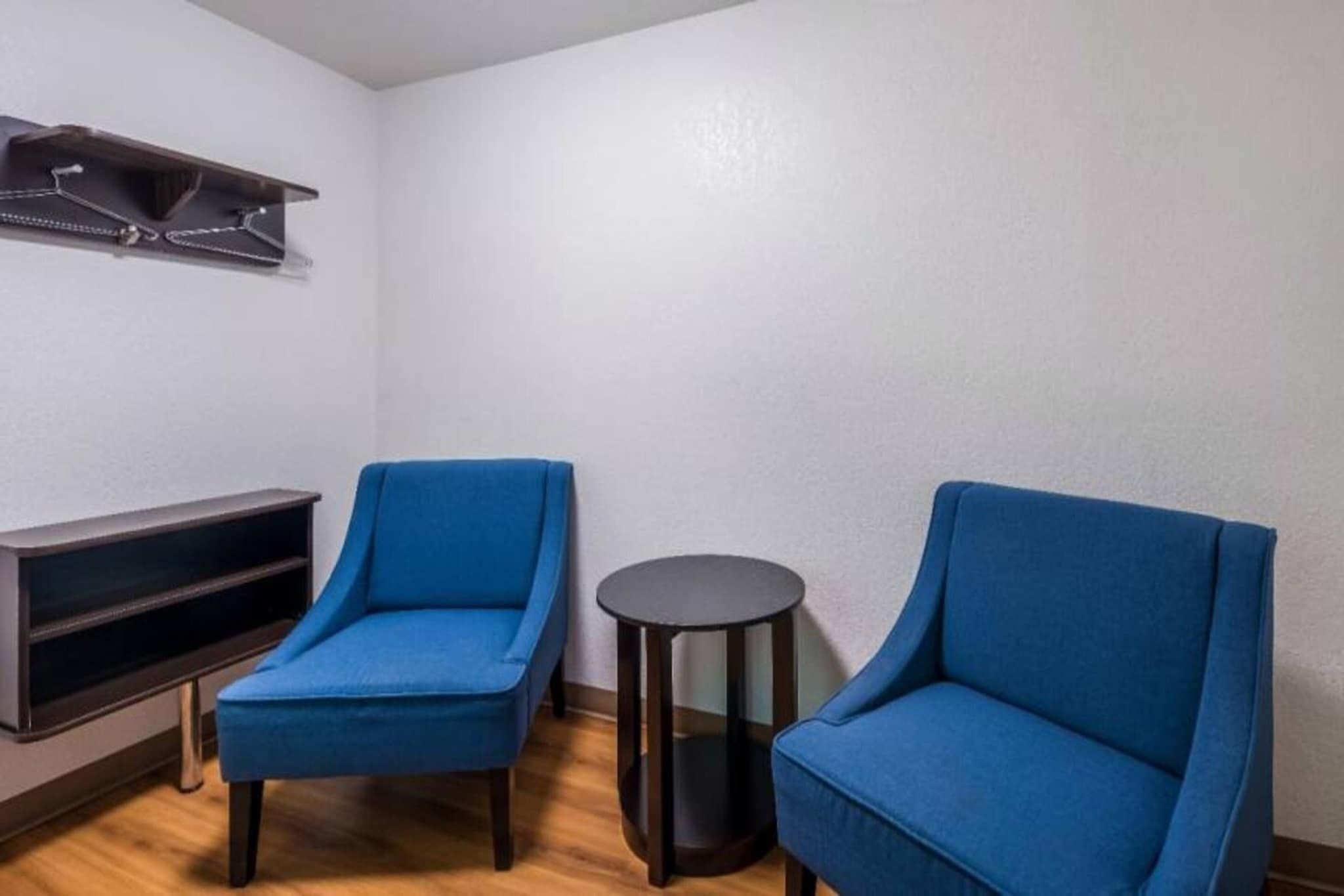 Una habitación con dos sillas azules, una mesita redonda y un estante oscuro contra la pared.