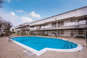 A rectangular swimming pool with a white border, surrounded by a metal fence, and a building with multiple balconies in the background. - 2 Queen Beds | Smoking, Micfridge (Irving TX)