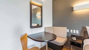 A compact room with a round table, two chairs, a sofa, a bedside table with a lamp, and a wall-mounted mirror.