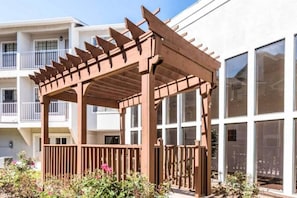 Eine hölzerne Pergola mit Balkon, umgeben von üppigem Grün und einem Gebäude mit großen Fenstern.