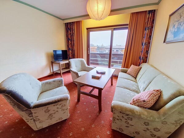 A hotel room with a sofa, armchair, TV, and a view of the city through the window. - Apartment Arnika by Interhome (Sankt Ulrich am Pillersee)