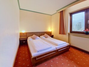A double bed with two pillows, a nightstand, a wall-mounted lamp, a window with curtains, and a radiator. - Apartment Arnika by Interhome (Sankt Ulrich am Pillersee)