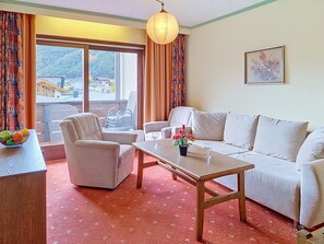 Ein Hotelzimmer mit einer Couch, Sesseln, einem Holztisch und Blick auf eine gebirgige Landschaft durch große Fenster.