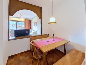 Uma sala de jantar com mesa de madeira, cadeiras, toalha de mesa rosa, luminária pendente e espelho refletindo um quarto com sofá e janela.
