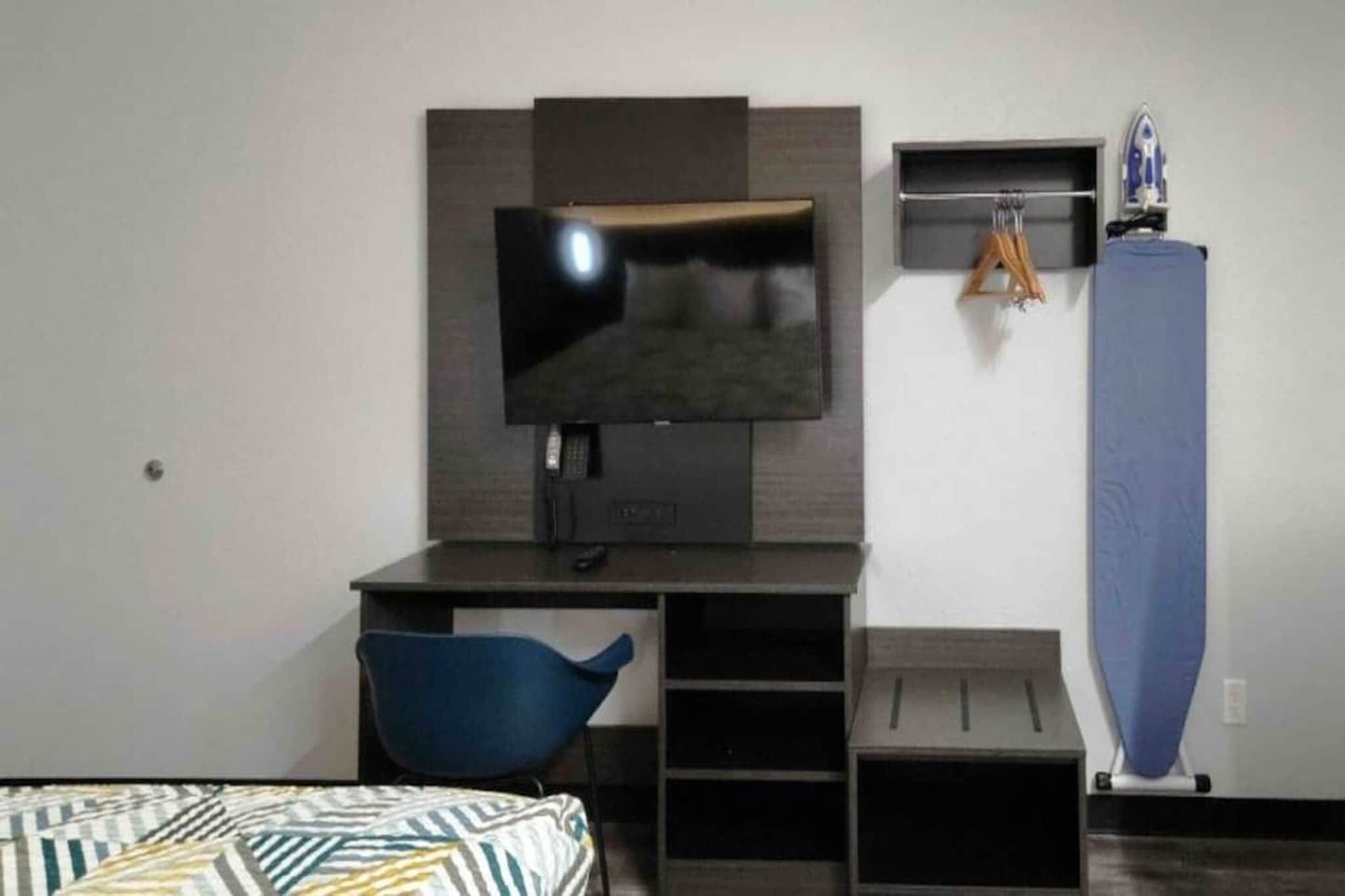A hotel room with a bed, a desk with a television, a blue chair, and a vertical ironing board.