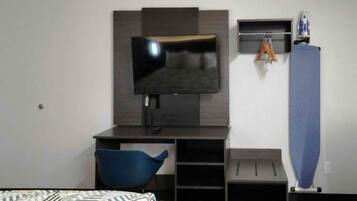 A hotel room with a bed, a desk with a television, a blue chair, and a vertical ironing board.