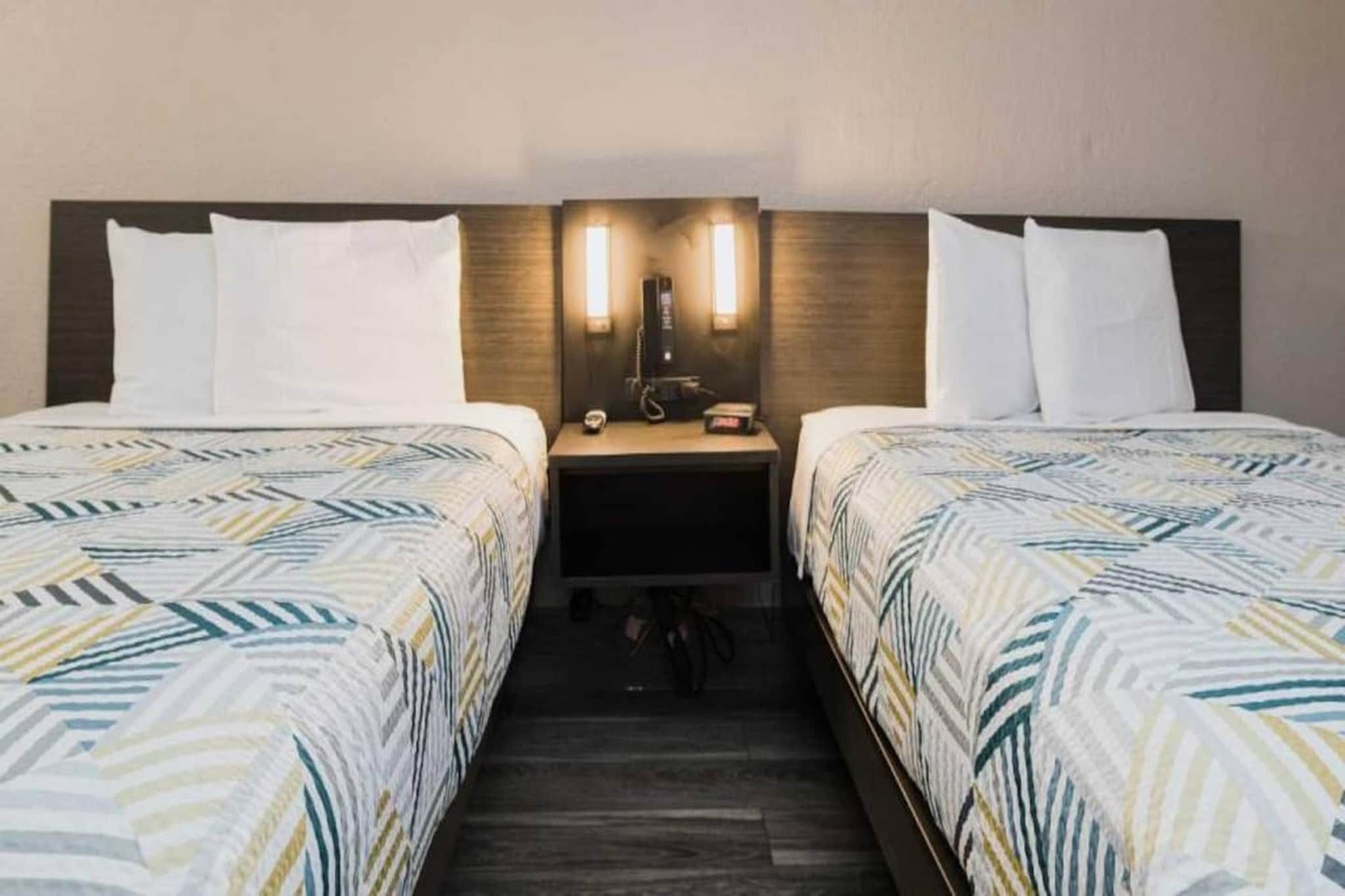 A hotel room with two beds, each with patterned bedding, a nightstand between them, and a wall-mounted light fixture.
