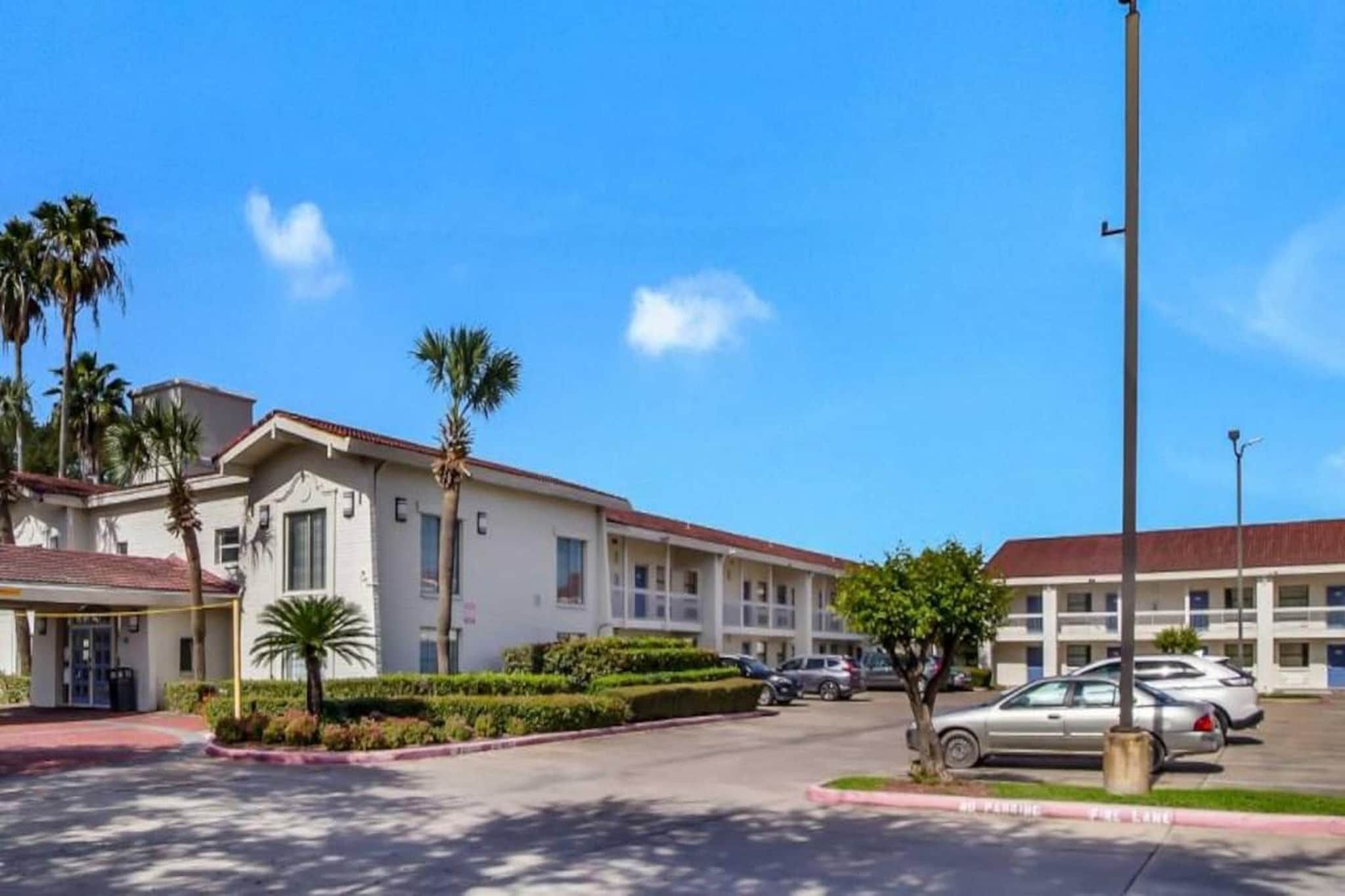 A hotel with a white facade, multiple stories, and a red roof. There are palm trees and a well-maintained lawn in front.