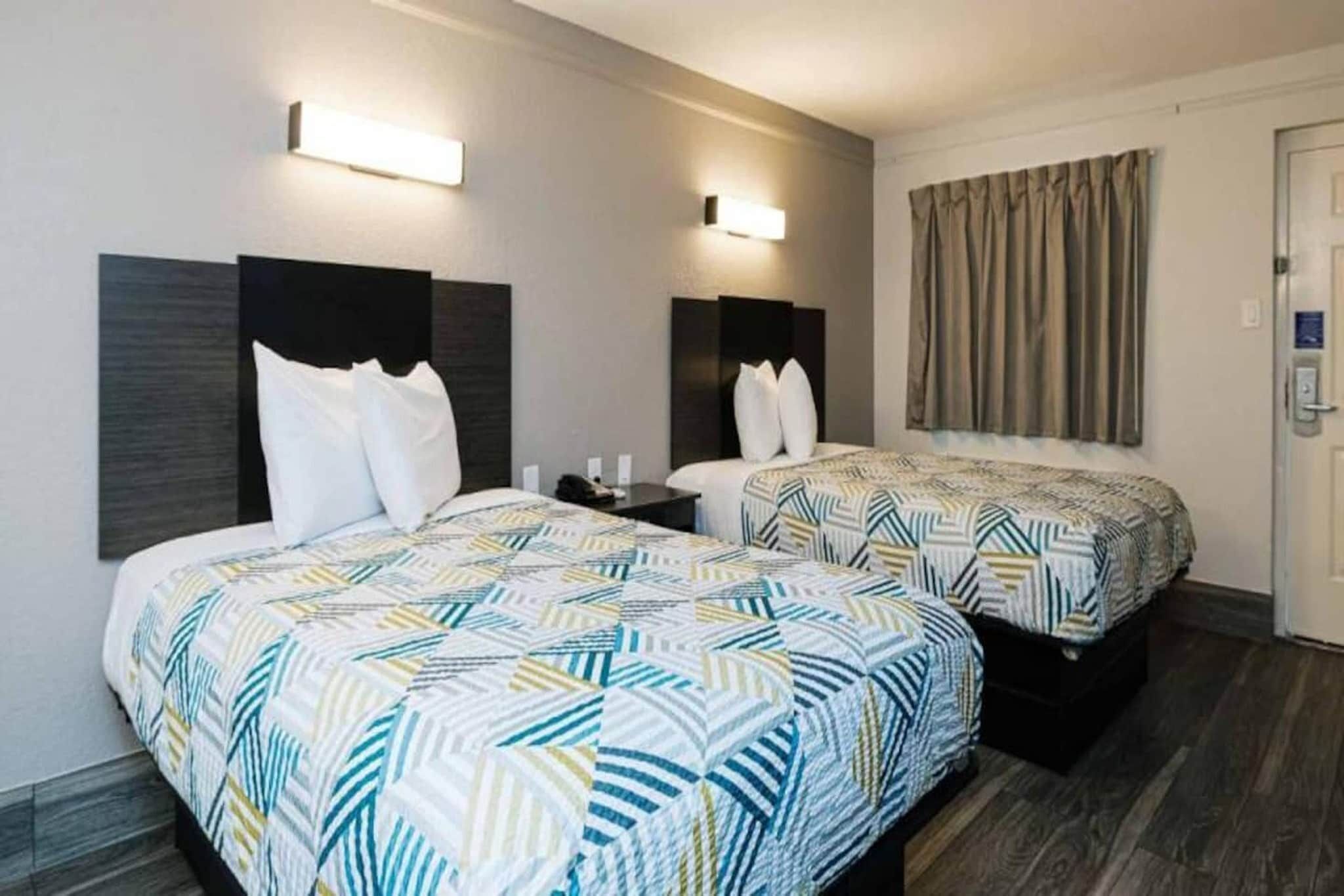 A hotel room with two beds, each with a patterned bedspread, a nightstand, a wall-mounted lamp, and a door with a curtain.