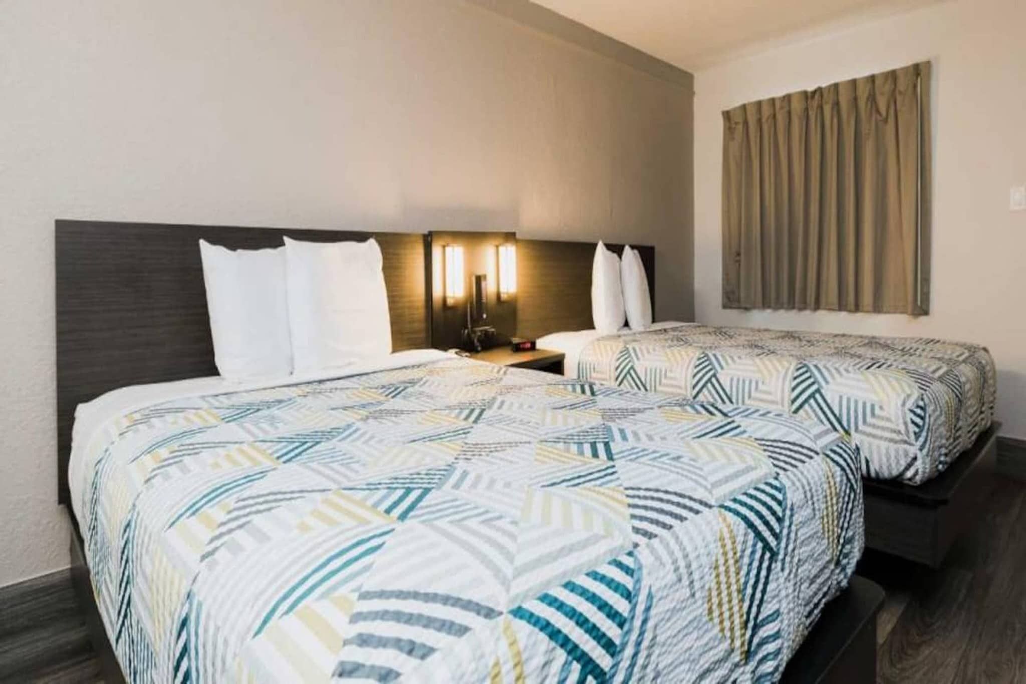 A hotel room with two beds, a nightstand, a lamp, and a window with curtains.