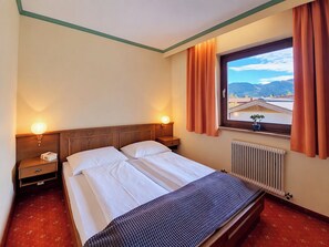 A hotel room with a bed, two bedside tables, a window with curtains, and a view of buildings and mountains. - Alpenrose by Interhome (Sankt Ulrich am Pillersee)