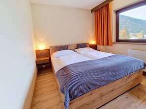 Ein Einzelbett mit Holz-Kopfstück, zwei Nachttische mit Lampen sowie ein Fenster mit Vorhängen.