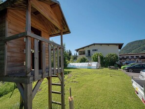 Eine Holzkonstruktion mit Balkon, einer Rasenfläche, einem Gewächshaus und einem Gebäude im Hintergrund mit einem Berg.