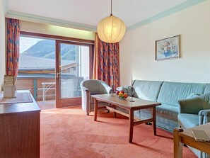 A hotel room with a sofa, a table with fruit, a chair, a desk, a lamp, a painting, and a view of mountains through the window. - Seerose by Interhome (Sankt Ulrich am Pillersee)