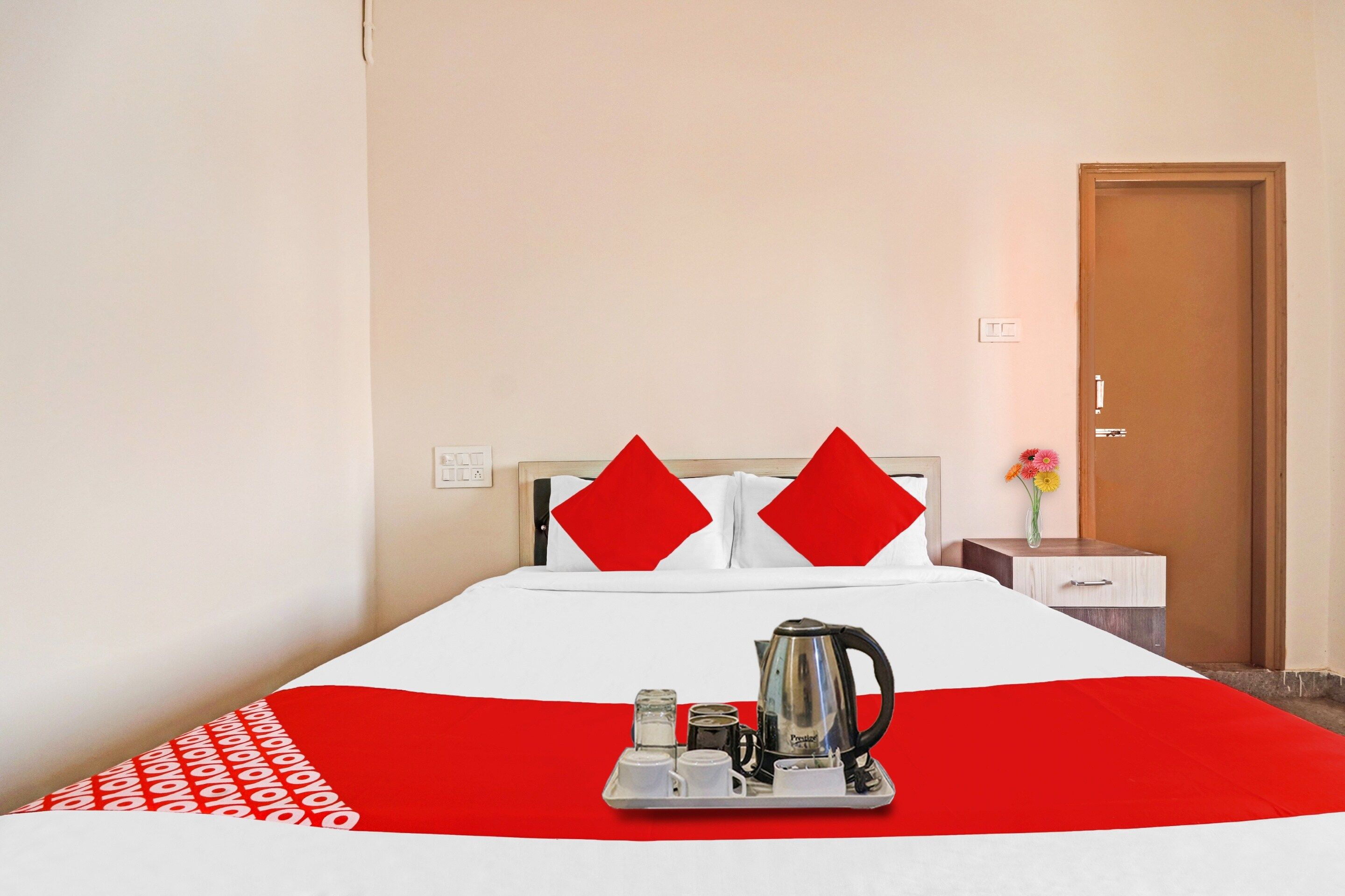 A hotel room with a bed, a nightstand, a vase with flowers, and a tray with a kettle and cups.