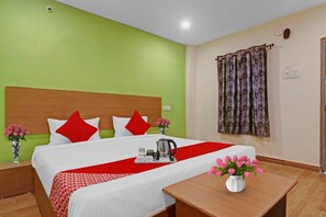 Quarto de hotel com cama, mesa de cabeceira, abajur, colcha vermelha e branca, um vaso com flores cor-de-rosa e uma janela com cortina.