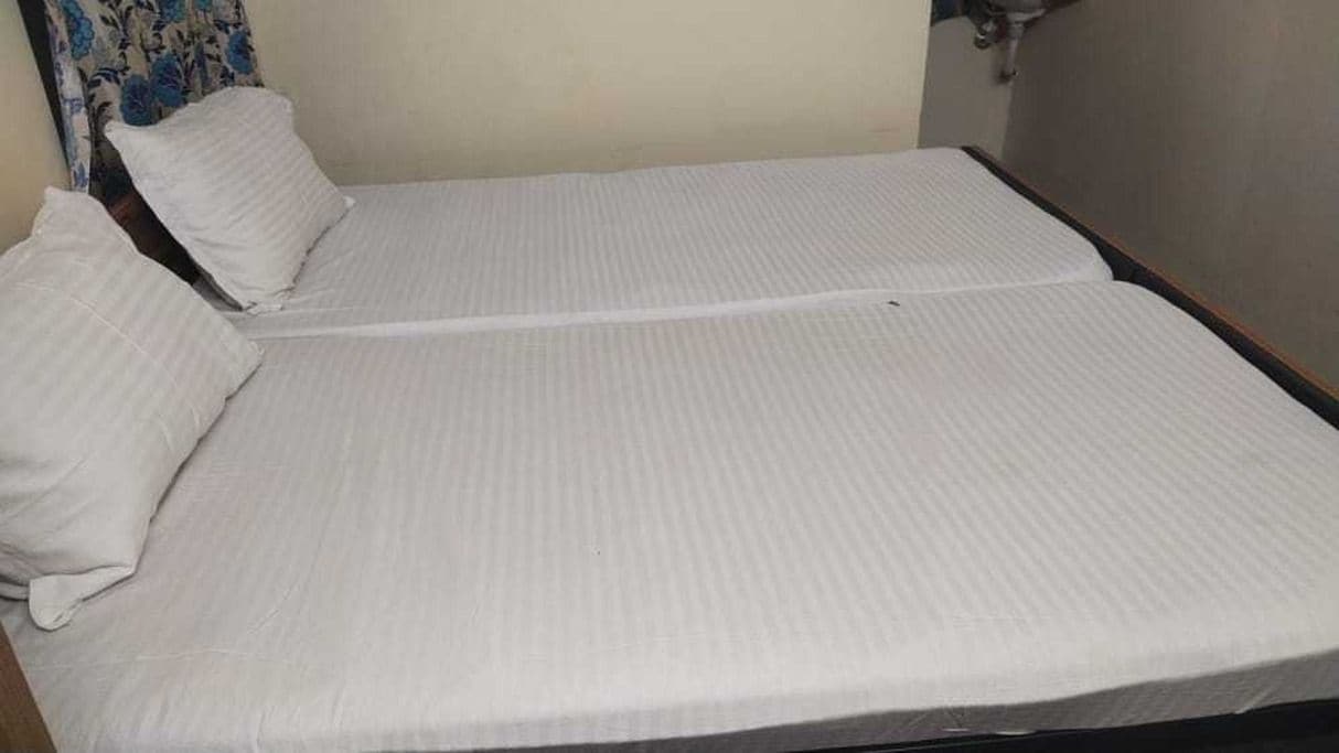 Una cama individual con colchón y almohadas blancos, cabecera de madera y una pared lisa con cortina.